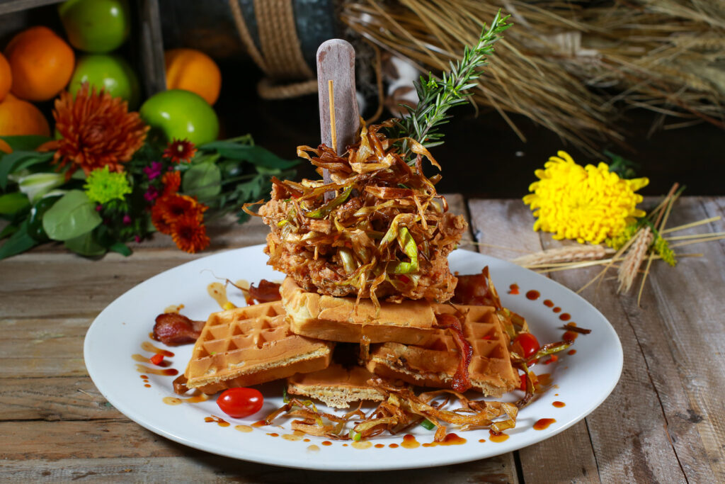 Towering fried chicken and waffles dish garnished with herbs and crispy onions at Hash House, served with syrup and cherry tomatoes in Las Vegas brunch setting.